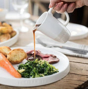 Person pouring gravy from a white pitcher onto a plate of food on a wooden table.