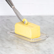 Butter knife spreading butter on a stick of butter on a glass dish with a marble background