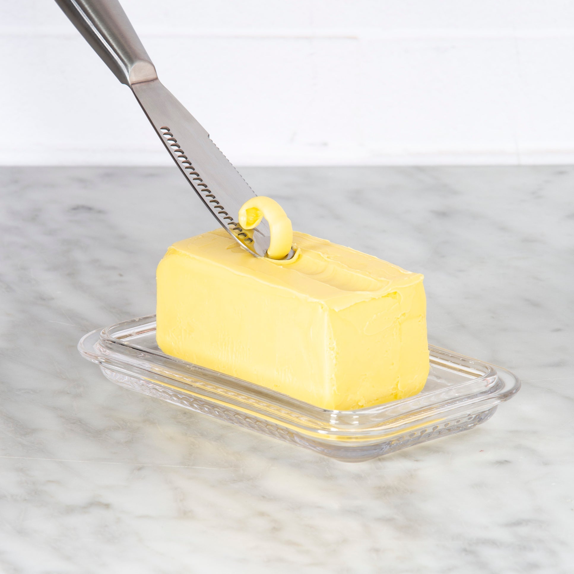 Butter knife spreading butter on a stick of butter on a glass dish with a marble background