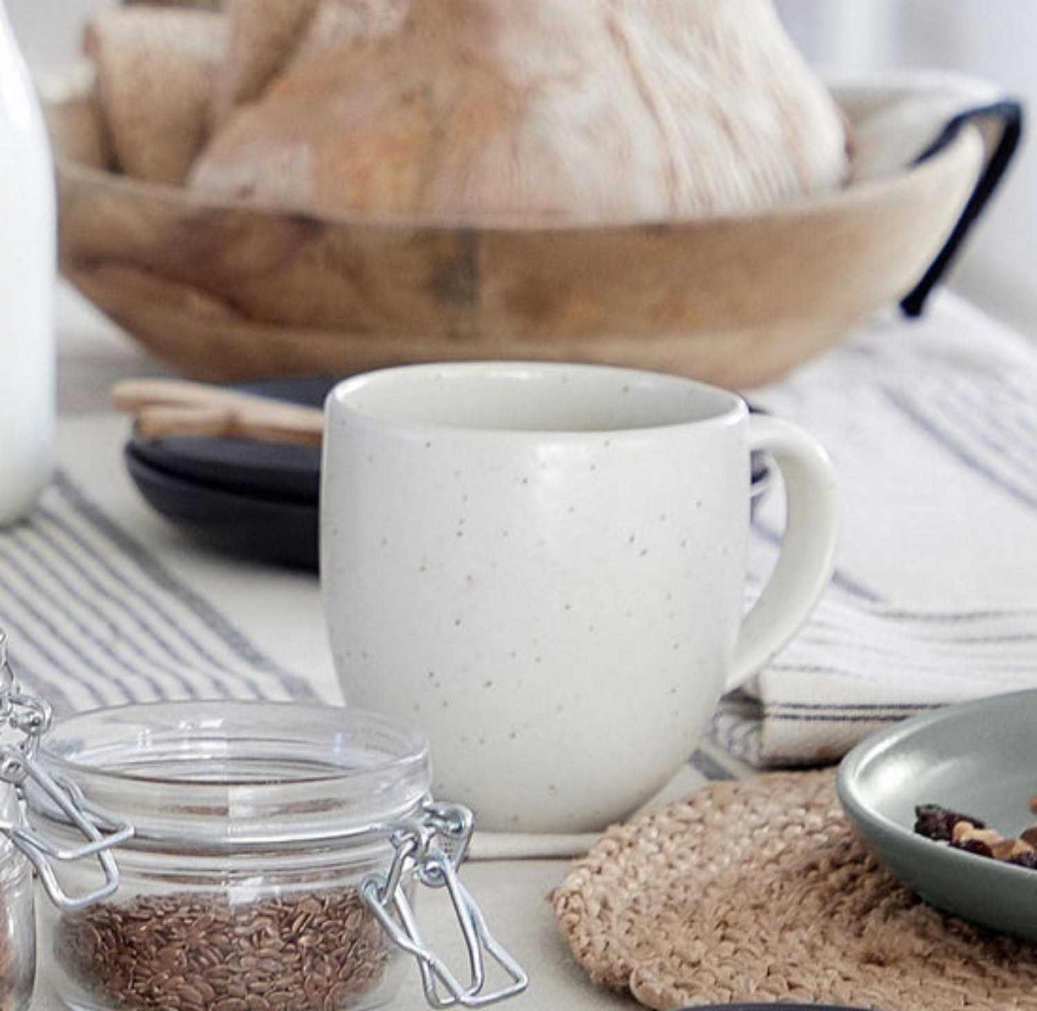 White speckled mug on a kitchen counter with a jar of coffee beans, a bowl of bread, and a striped towel.