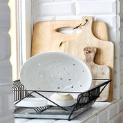 White ceramic strainer with holes, wooden cutting board, and black metal tray on a white tiled wall background