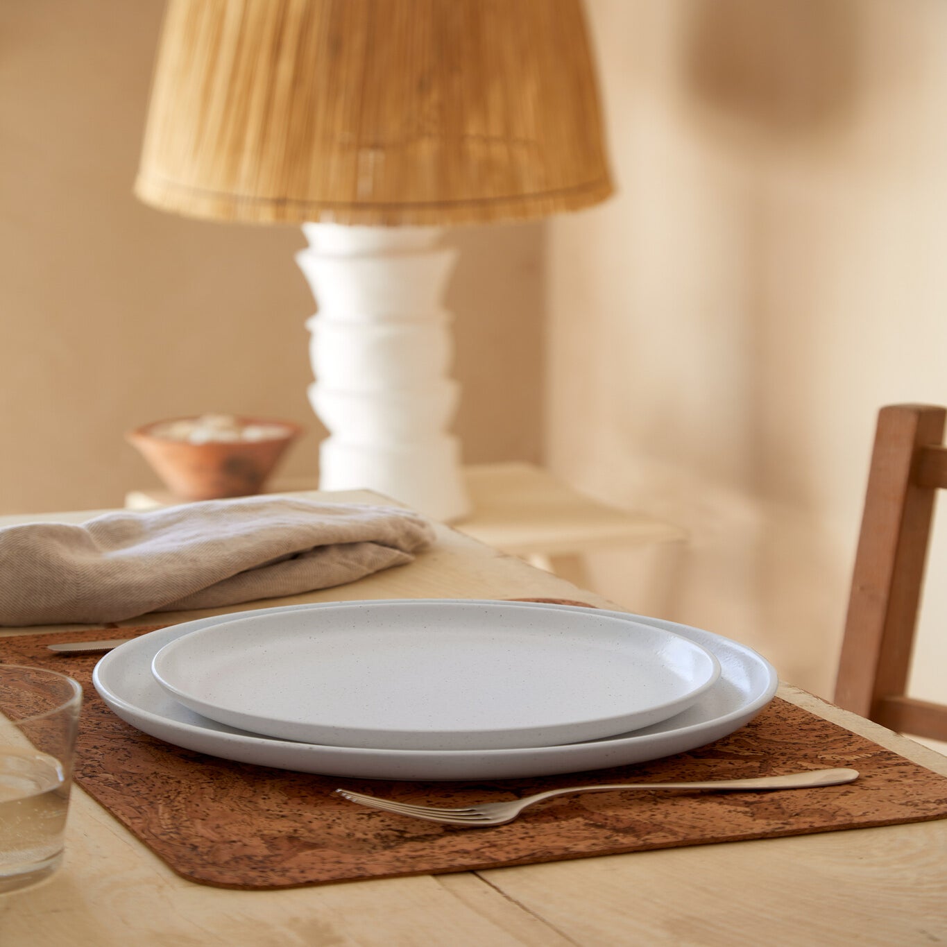 Dining table setting with plates, placemat, and lamp in a warm-toned room.