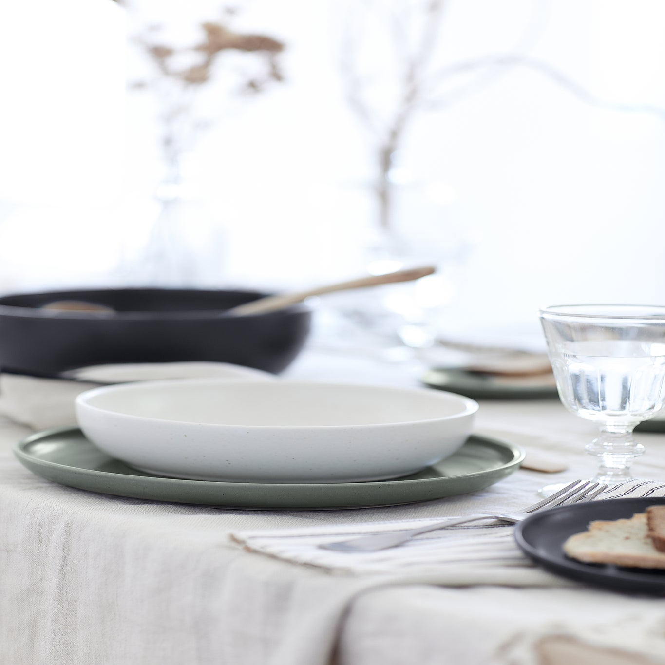 Dining table setting with white bowl, green plates, and glass on a neutral background