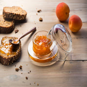 Glass serving dish of apricot jam with a spoon, sliced bread, and apricots on a wooden surface.