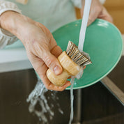 Person washing a wooden dish brush under running water with a green plate in the background.