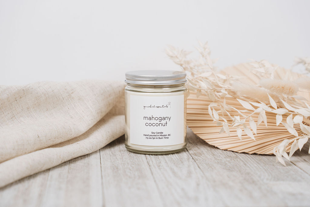 Candle labeled 'mahogany coconut' on a wooden surface with a beige blanket and dried flowers.
