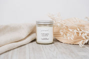 Candle labeled 'mahogany coconut' on a wooden surface with a beige blanket and dried flowers.