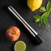 Black and silver cocktail muddler with a lemon, peach, and lime on a dark surface