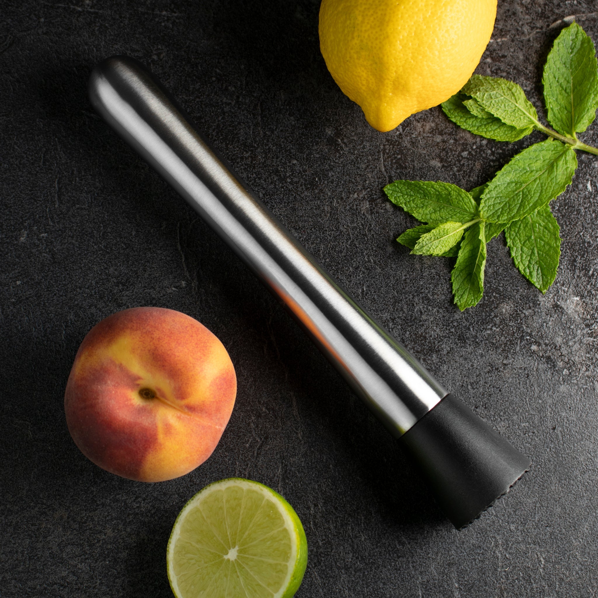 Black and silver cocktail muddler with a lemon, peach, and lime on a dark surface