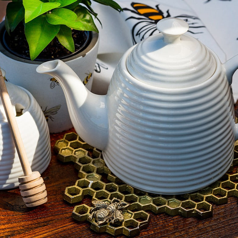 glossy white bone china tea pot in shape of beehive with long curled spout sitting on gold honeycomb trivet on dark wood table. Various bee themed items in background with green plant, honey pot with wood honey dipper 
