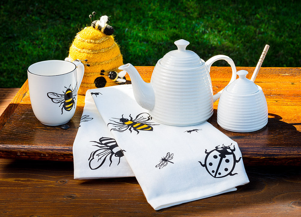 glossy white bone china tea pot in shape of beehive with long curled spout beside glossy white honey pot with dipper, white mug with yellow honey bee, felt beehive and bee tea towel on dark wood table with green grass in background 
