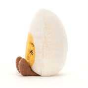 side view of fuzzy white and yellow boiled egg stuffed plush toy with black squinting smiley face made by jellycat