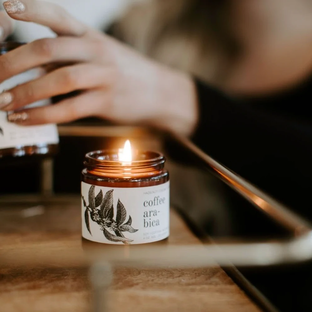 lit coffee arabica candle brown jar white label black lid hand in background blurred