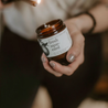 fresh squeezed candle brown jar white label black lid being held by hand and lit body in background blurry