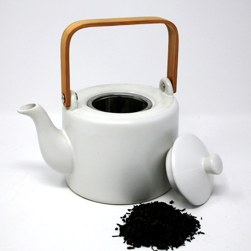 matte white teapot with wood handle and metal strainer. Open top with lid propped up against  side. small pile of black loose leaf tea in front. On white background