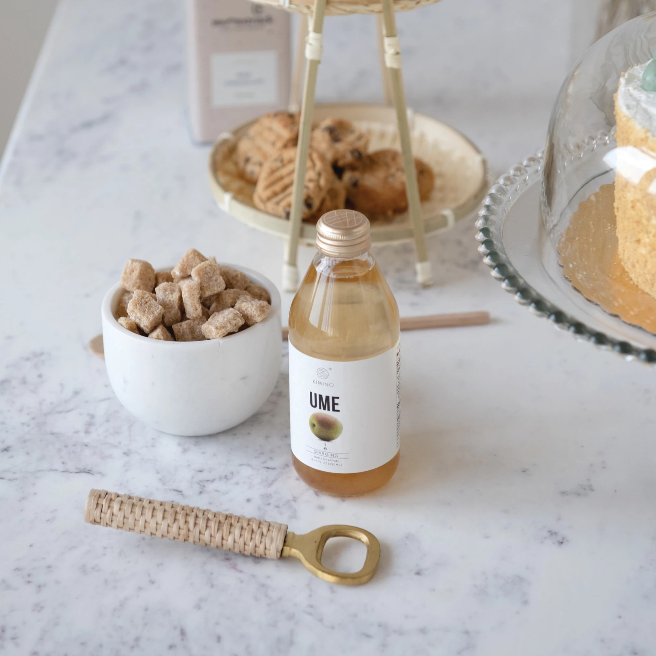 marble counter with plate with cookies and cake on background. in front is a bowl with sugar chunks, a bottle of ume and gold and rattan wrapped bottle opener 