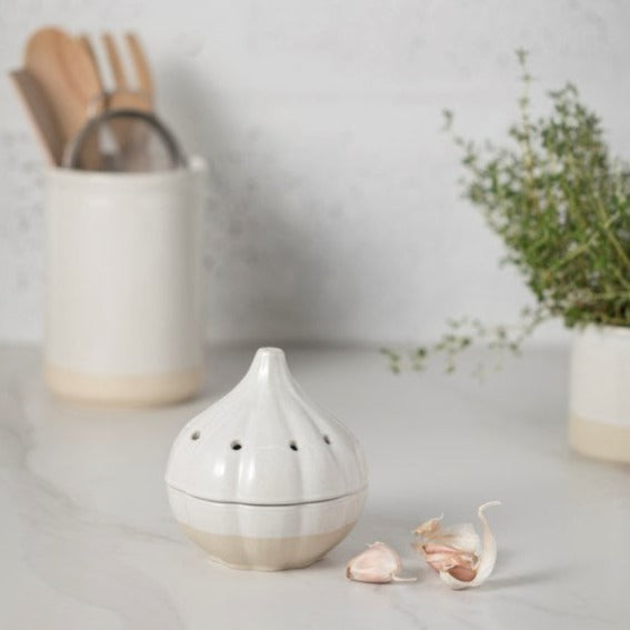 two tone stoneware garlic keeper with loose garlic beside on countertop with utensils behind and pot of herbs 
