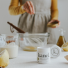 white tube black writing of charred lemon sea salt in foreground in background out of focus a white person in gray apron yellow sleeves sprinkling salt into a glass bowl with whisk oil and lemons at side 
