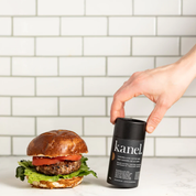 side view of burger on left beside on right is a white hand holding a tube of caramelized coffee rub on angle in background is out of focus subway tile backsplash