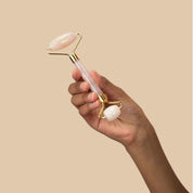rose quartz and gold facial roller held by black hand in air, middle of frame with tan background behind