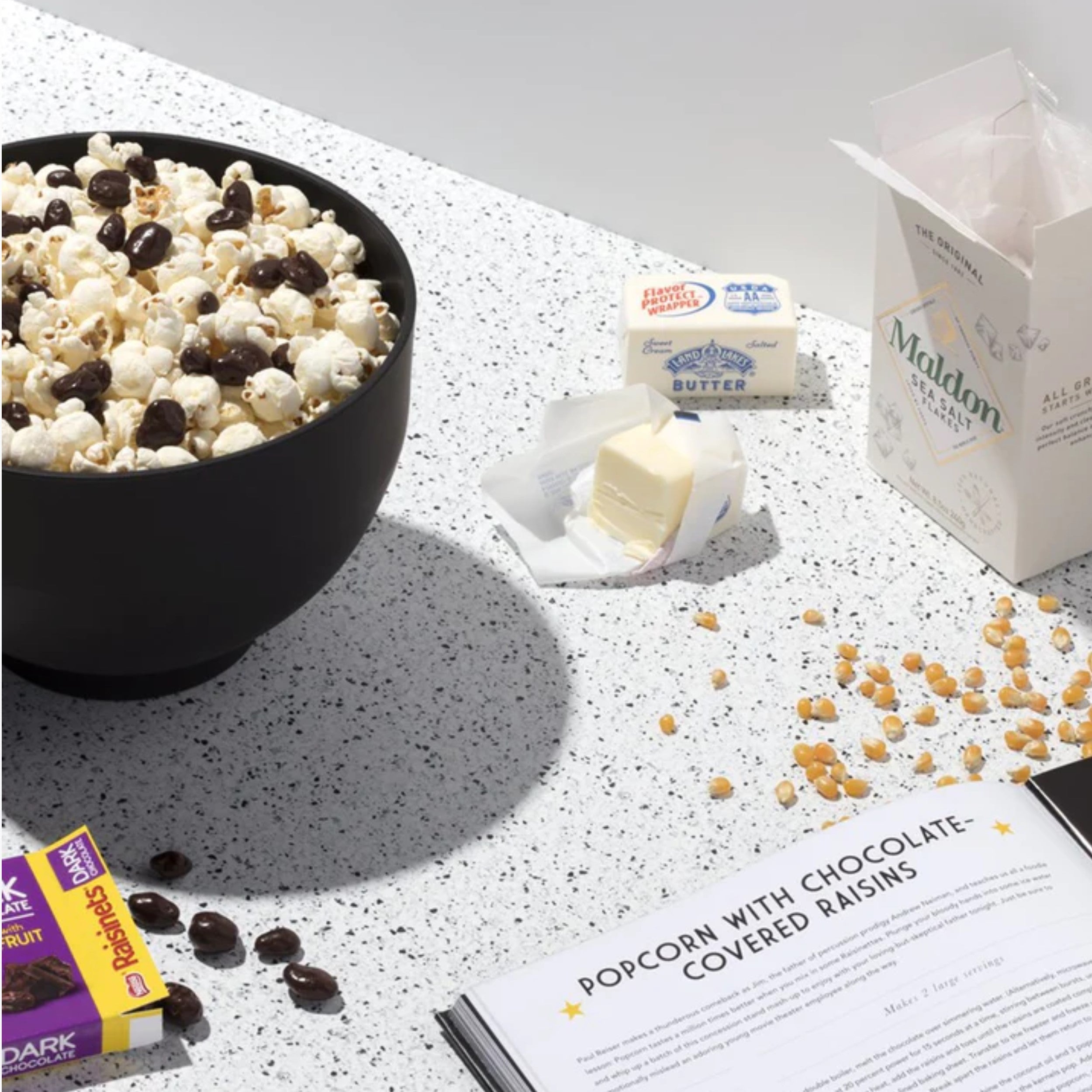 white speckled counter with silicone bowl of popcorn and chocolate raisins at top left corner, below is corner of purple and yellow box of chocolate raisins, right side top is box of maldon salt  and two butter bricks, below loose popcorn kernels and at bottom is a cook book open with a recipe  